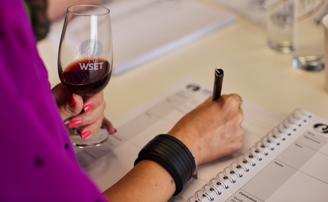 A person holding a tasting glass with some red wine, writing a tasting note.